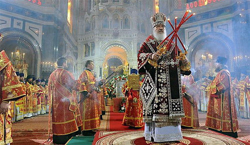 Orthodox faithful standing in prayer during the Divine Liturgy, holding candles in a church filled with incense
